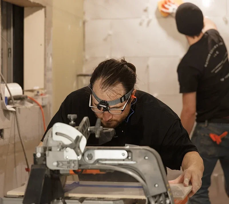 Two construction workers, one cutting tiles on a machine, the other applying grout to a tiled wall.
