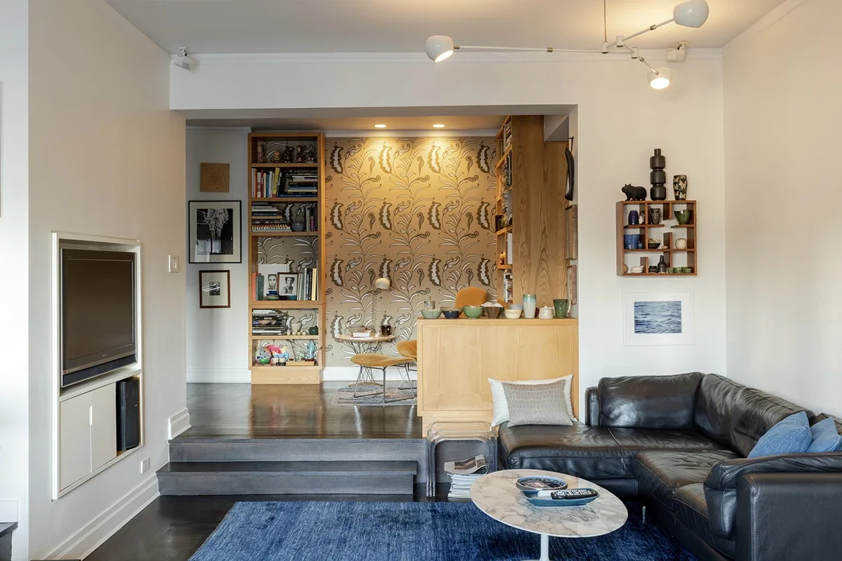 A living room with a black sectional, blue rug, and marble coffee table. Beyond, a raised dining area with floral wallpaper.