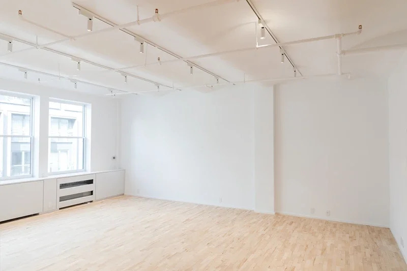 A bright, empty room with white walls, light wood floors, track lighting, and two large windows on the left wall.