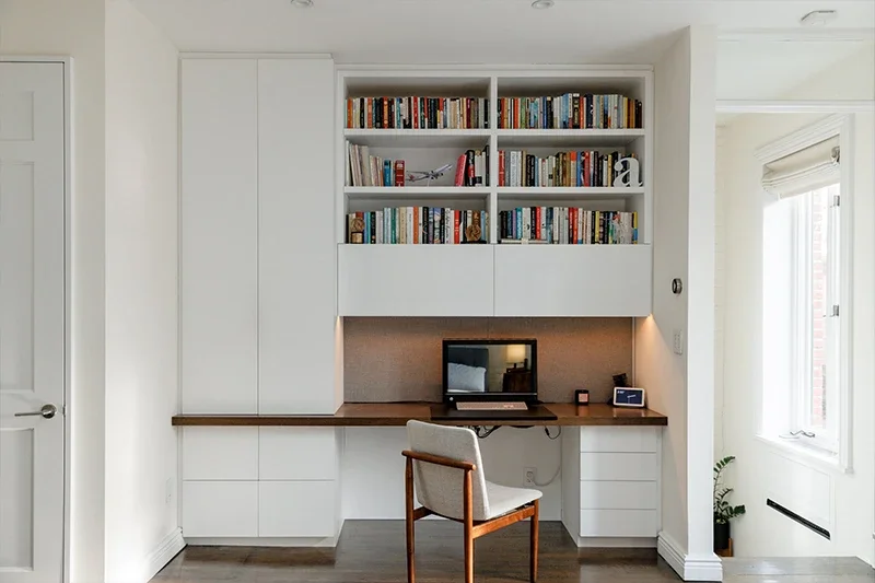 A modern home office with a built-in white desk, bookshelves, storage, a laptop, and a beige chair.