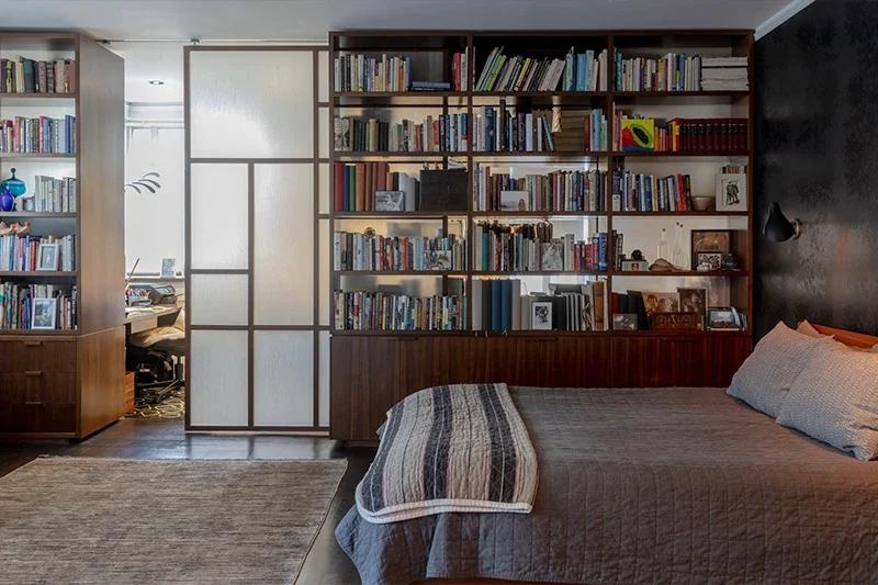 A bedroom with a bed and a large built-in bookshelf, next to a translucent sliding door that leads to an office.