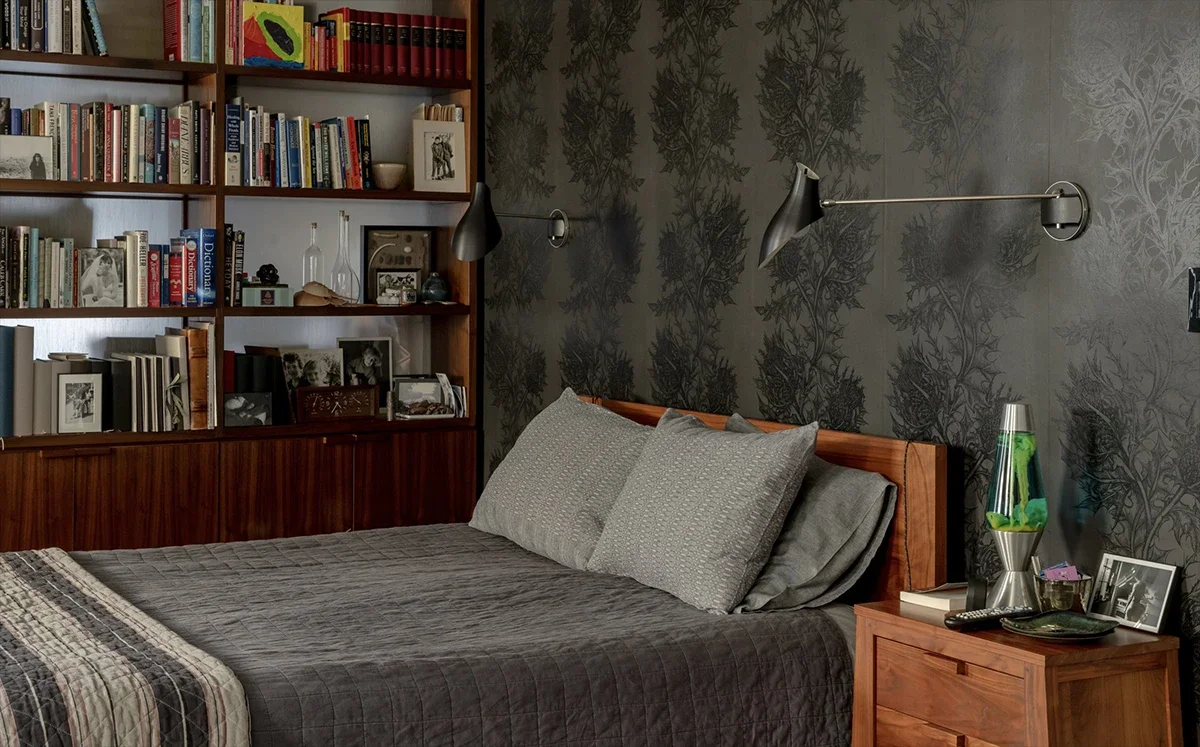 A dark bedroom with patterned wallpaper, a made bed with gray quilts, a wooden headboard, and a large bookshelf.