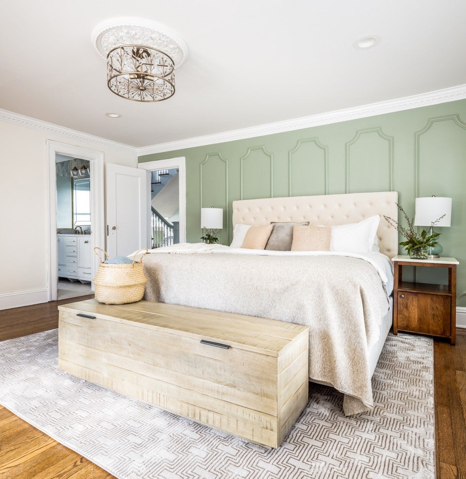 A beige upholstered bed with patterned pillows sits on a patterned rug in a stylish green room.