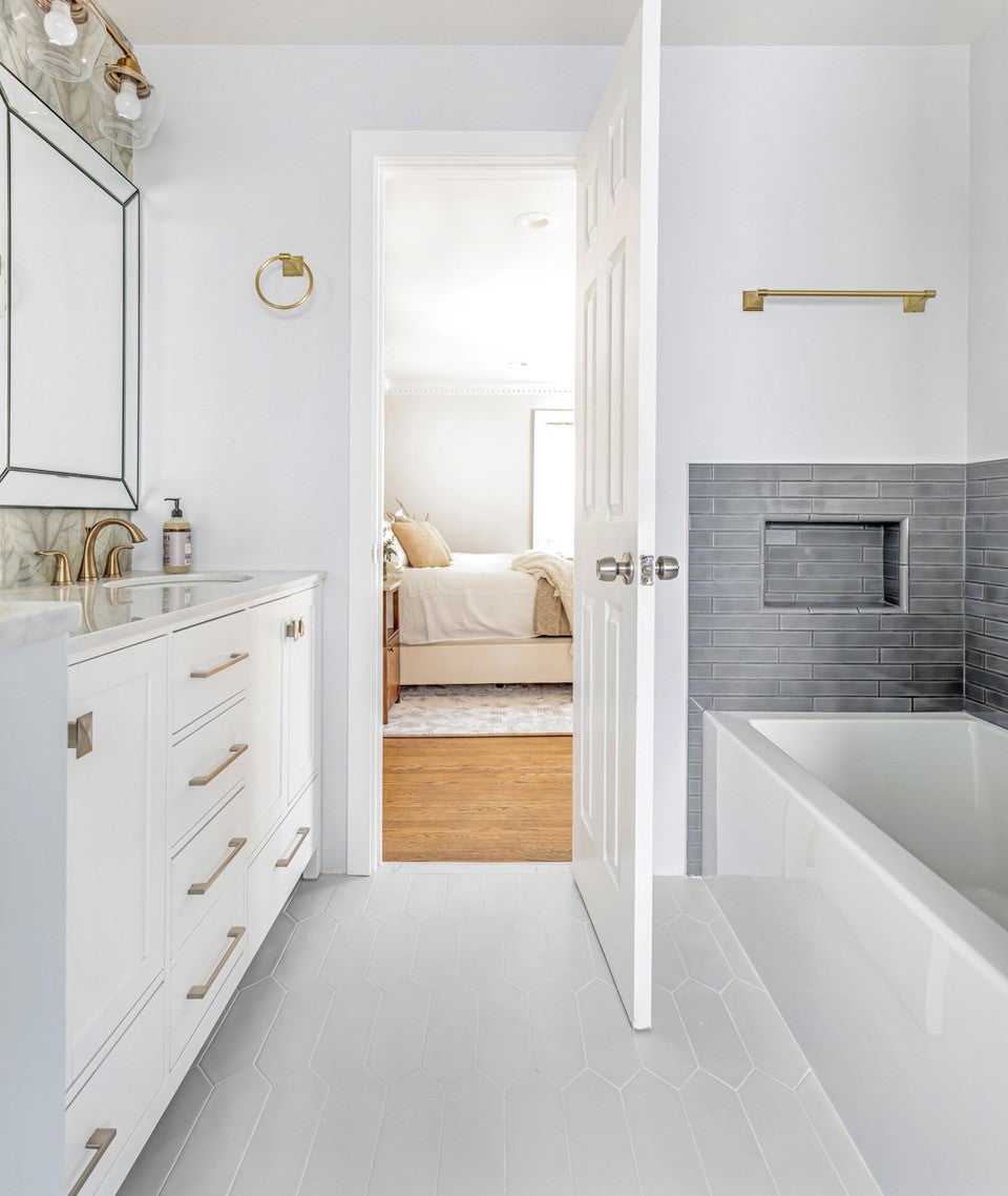 A white bathroom with gray tiled accents opens into a bedroom with a bed and hardwood floors.