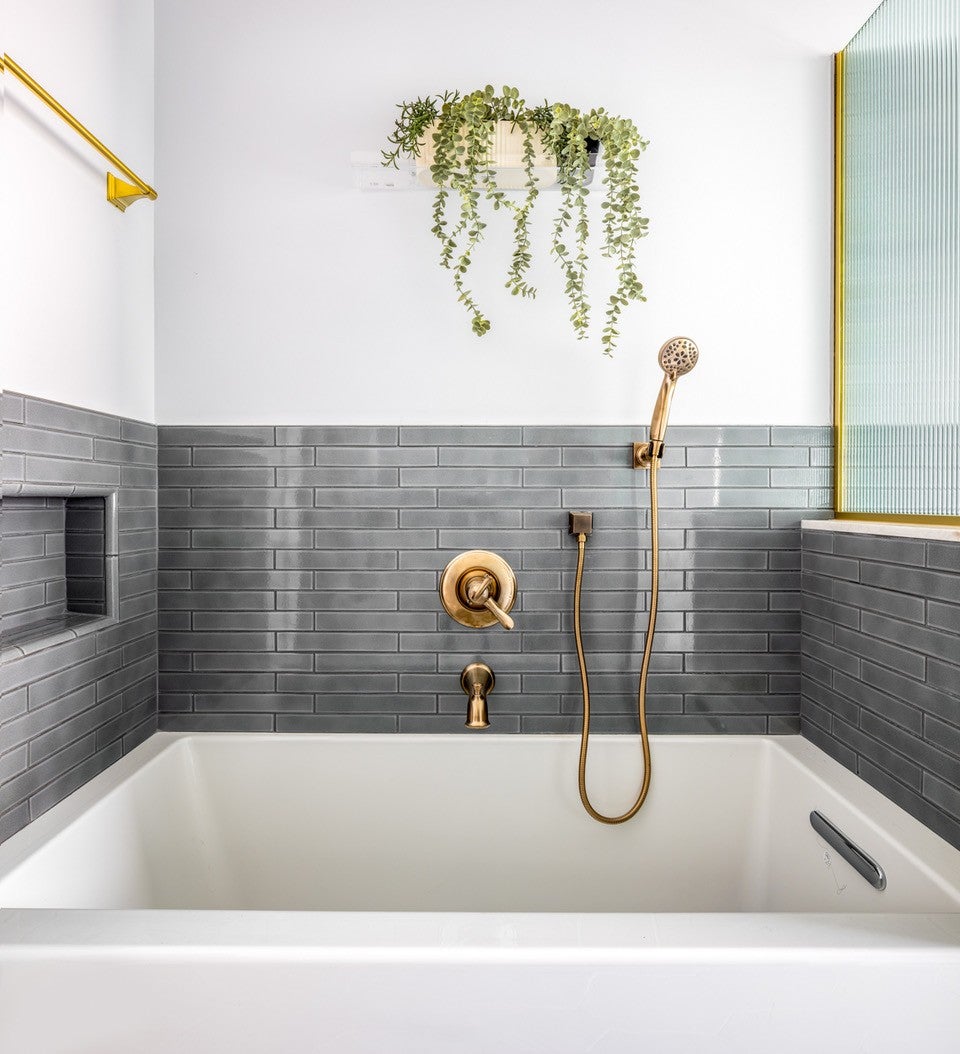 A modern bathroom features a white bathtub, gray tiled walls, and gold-toned shower fixtures and decorative hanging plant.