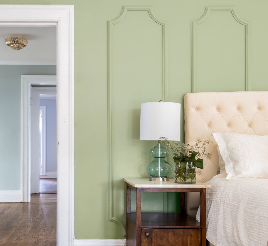 A bedside table with a lamp and flowers sits next to a tufted headboard in a green-walled room.