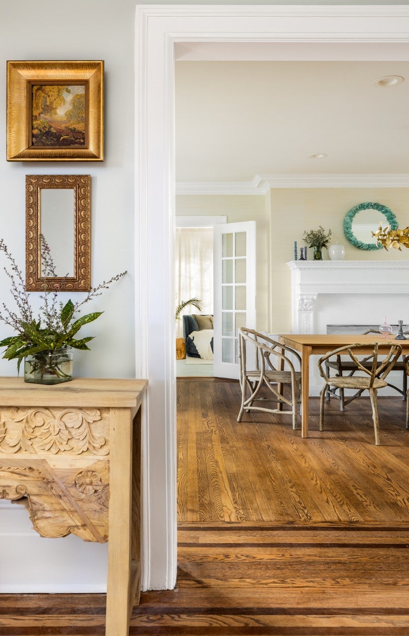 A vintage-framed painting hangs above a carved wooden console table in a bright, open interior.