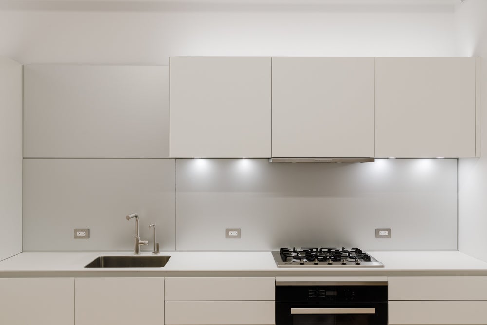 A modern kitchen with light gray cabinets and white counters. A sink is on the left, and a stovetop and oven are on the right.