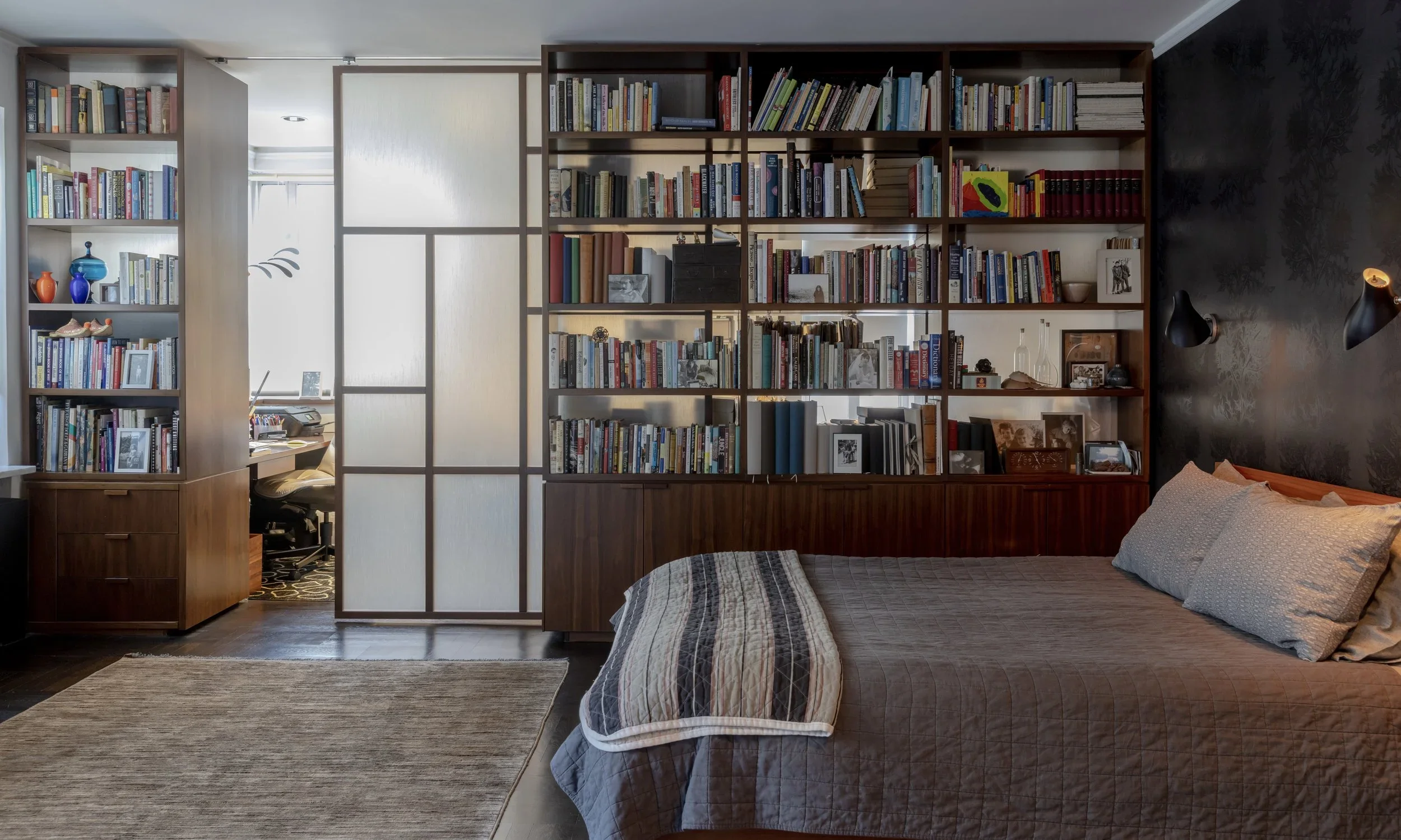 A bedroom with a dark grey patterned wallpaper, a dark wood bookcase, and a bed with grey bedding.