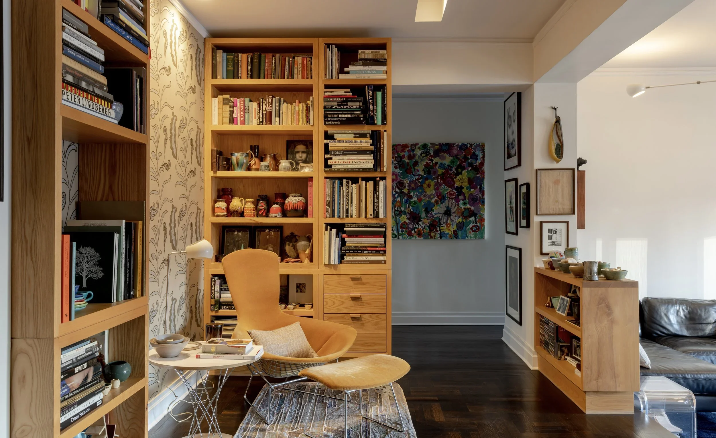 A cozy reading nook with a mustard yellow armchair and ottoman in front of a tall wooden bookshelf filled with books.