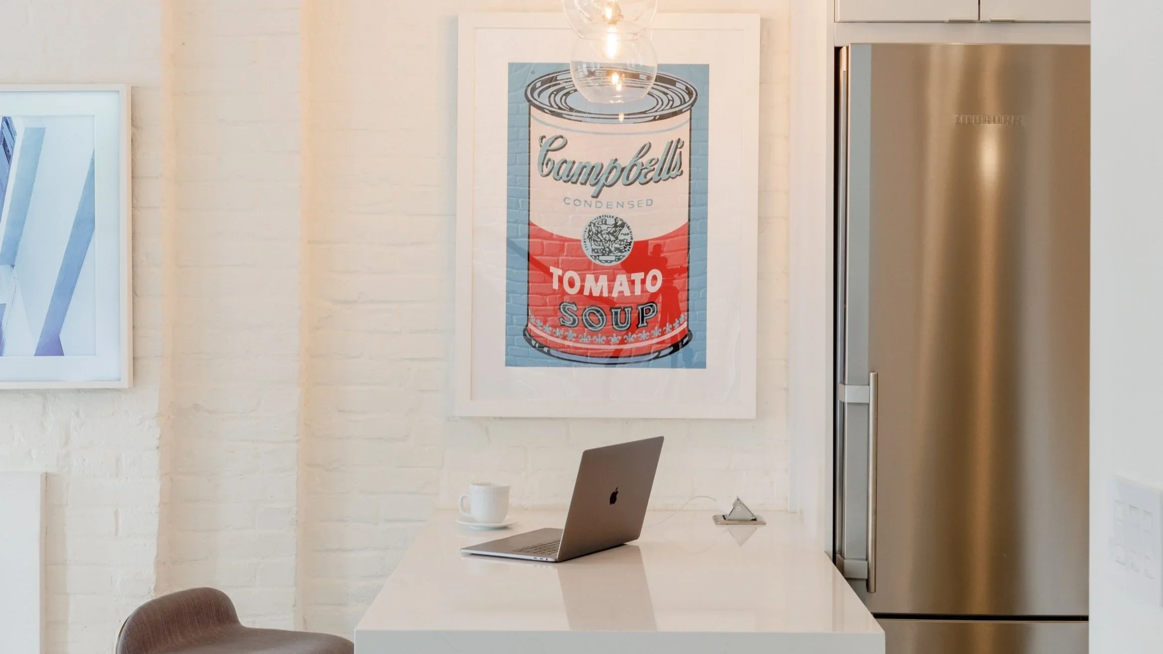 Modern kitchen workspace with white counter, MacBook, Andy Warhol soup painting, and stainless steel fridge.