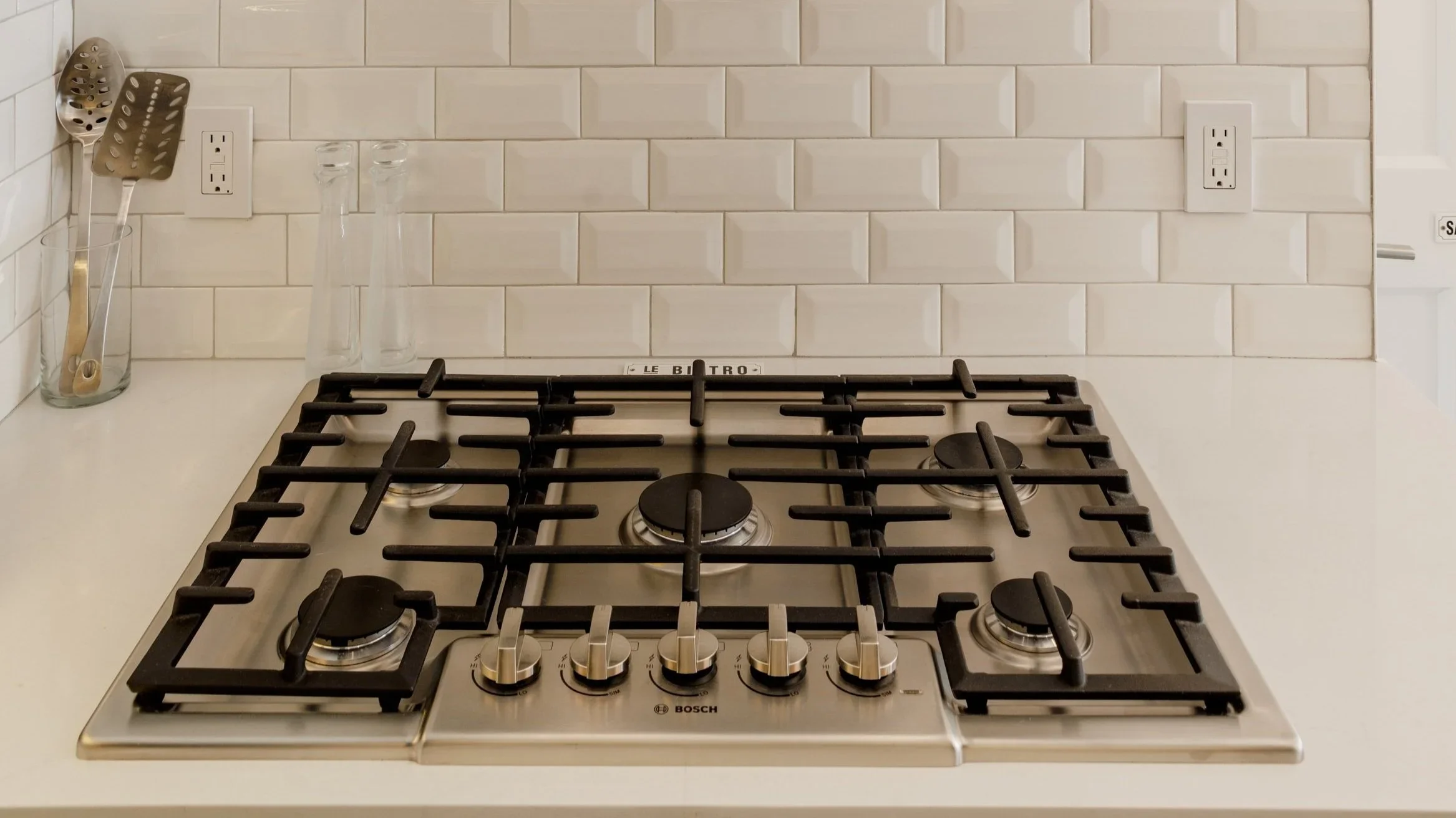 Stainless steel gas stovetop with black grates and five burners, set into a white counter, with white subway tile backsplash.