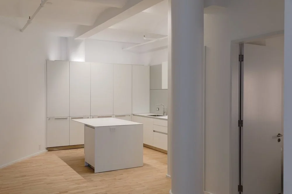 Minimalist white kitchen with a freestanding island and light wood flooring, viewed from a doorway.